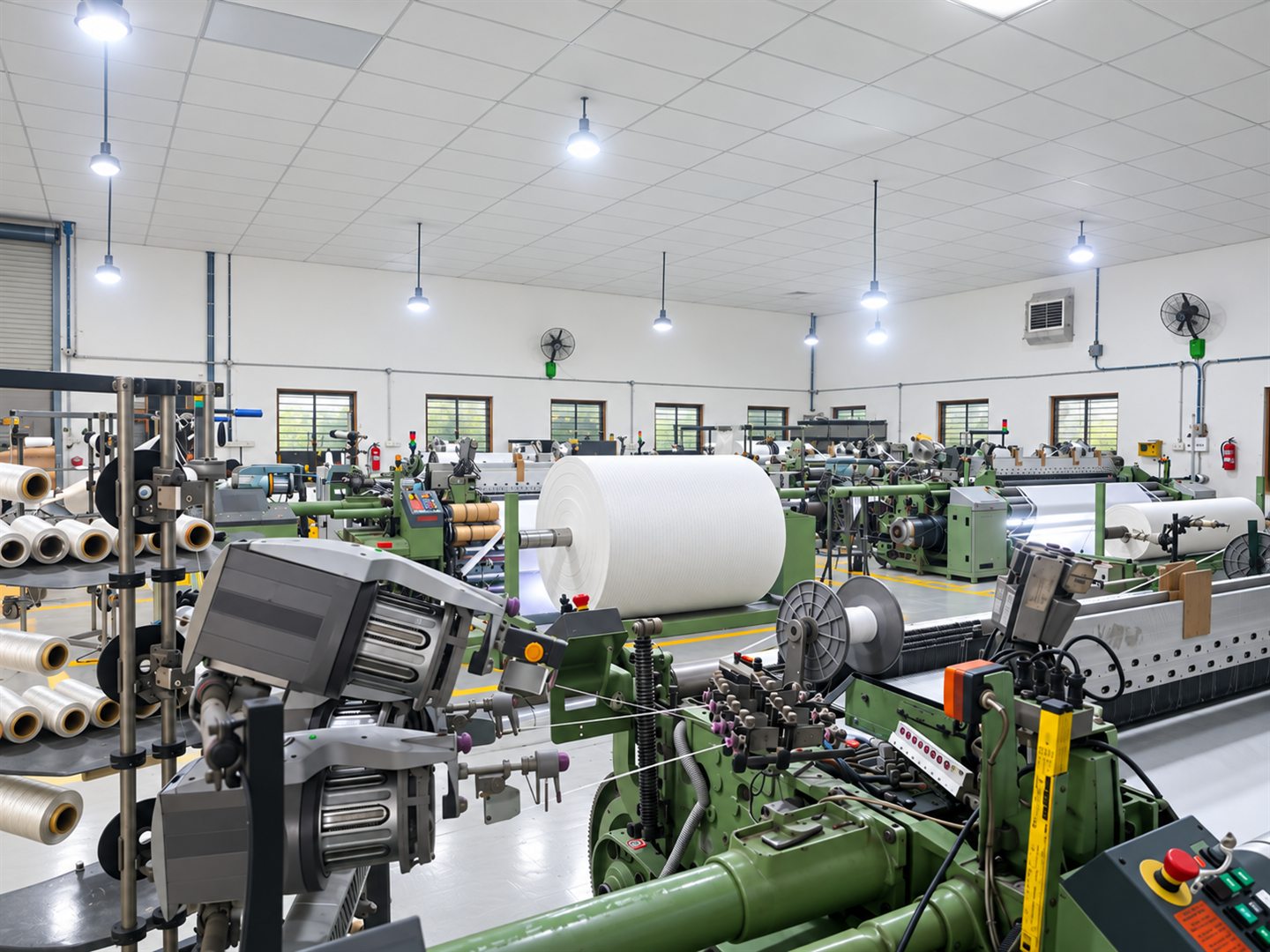 The DFR weaving floor at Unit-1 — rapier looms running either side of a full roll of woven filtration fabric, with the yarn creel racked along the left wall